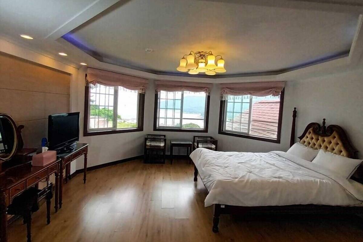 A hotel room with a bed, a television, a desk, and a chair. There are windows with curtains, a ceiling light, and a view of the ocean.