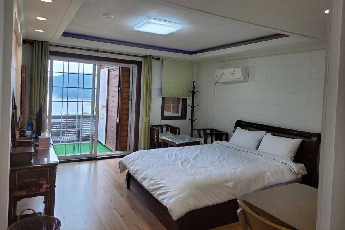 A hotel room with a bed, a desk, a chair, a small table, and a view of the sea through a sliding door.