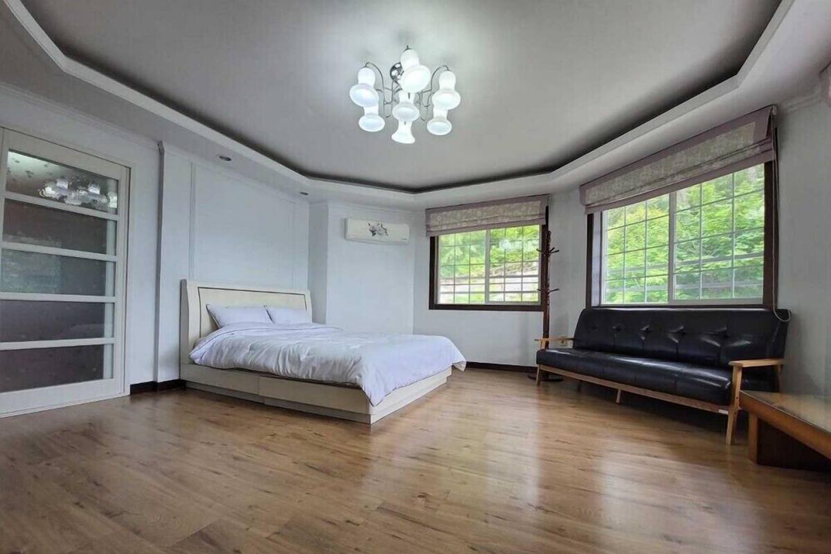 A bedroom with a bed, a black leather sofa, a chandelier, and large windows.