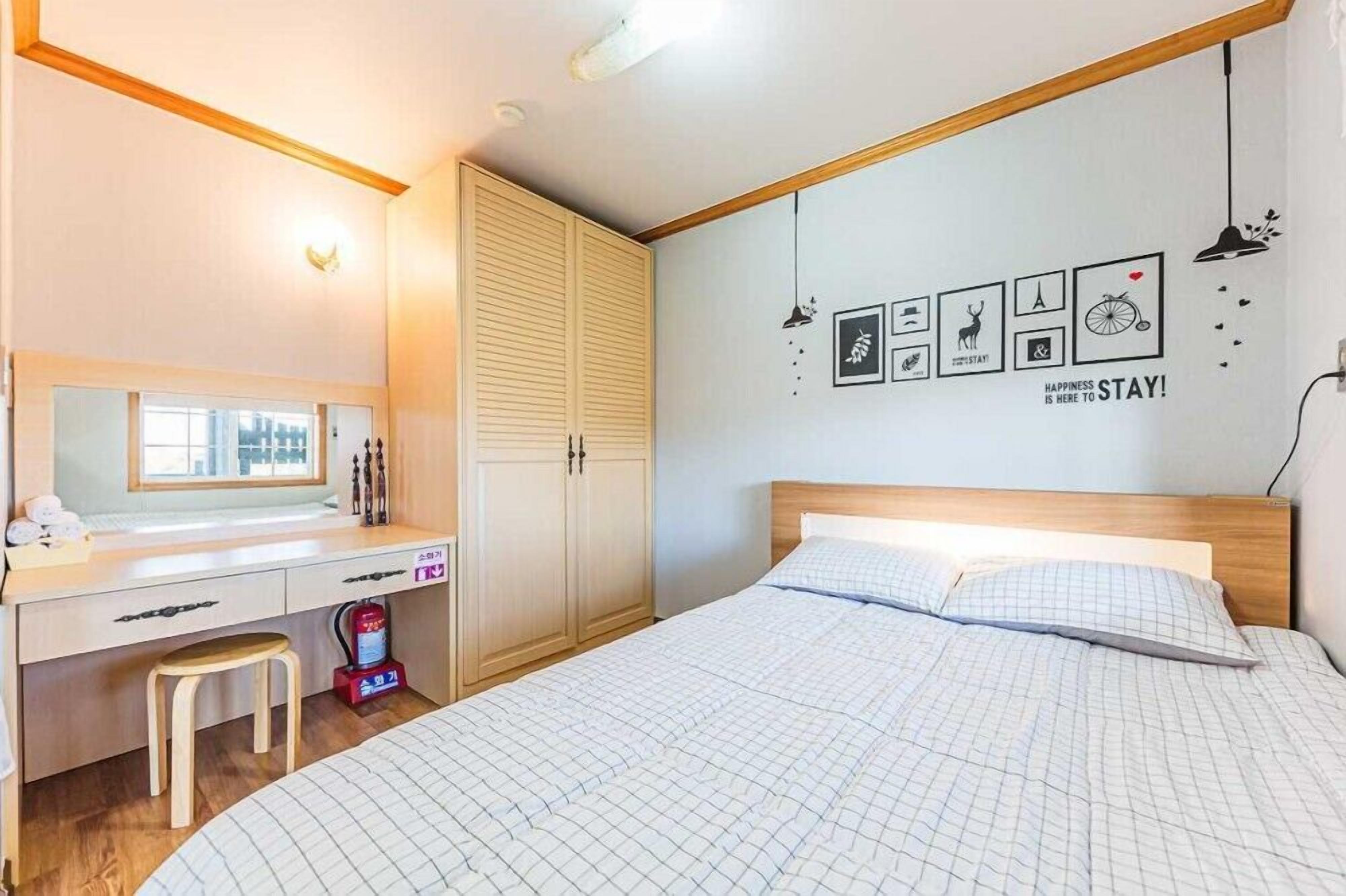 A bedroom with a bed, a desk, a chair, a wardrobe, and a wall with framed pictures and a motivational quote.