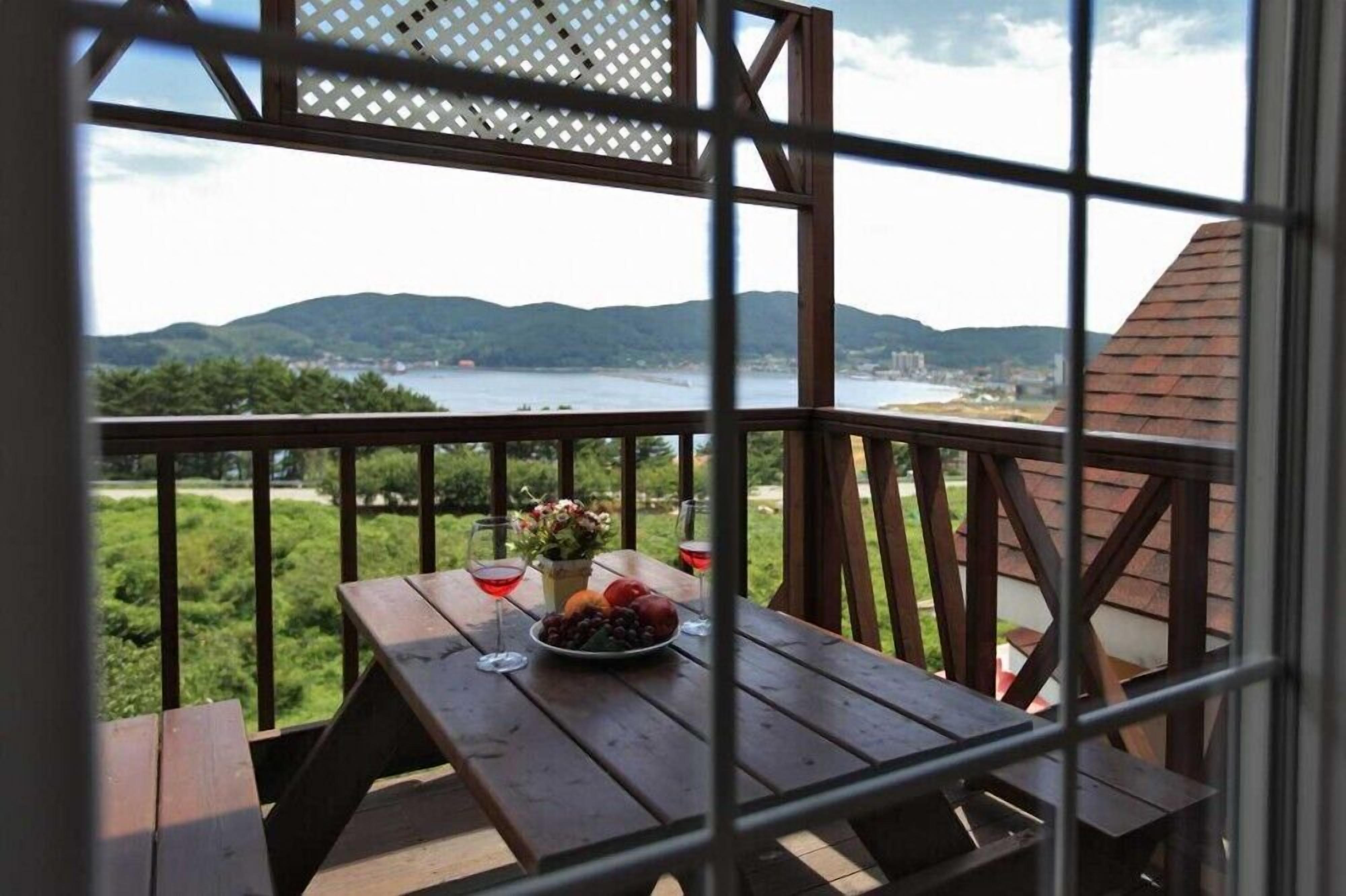 Una mesa de madera con un frutero y copas de vino en un balcón con vistas a un lago y colinas.