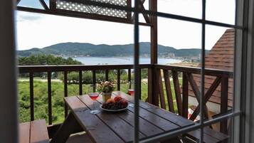 Una mesa de madera con un frutero y copas de vino en un balcón con vistas a un lago y colinas.