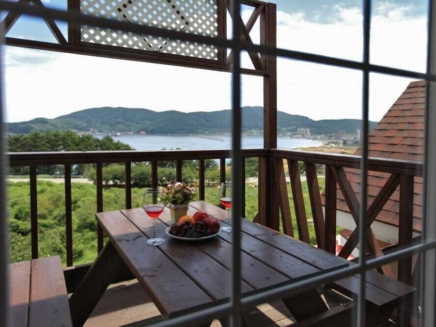 Una mesa de madera con un frutero y copas de vino en un balcón con vistas a un lago y colinas.