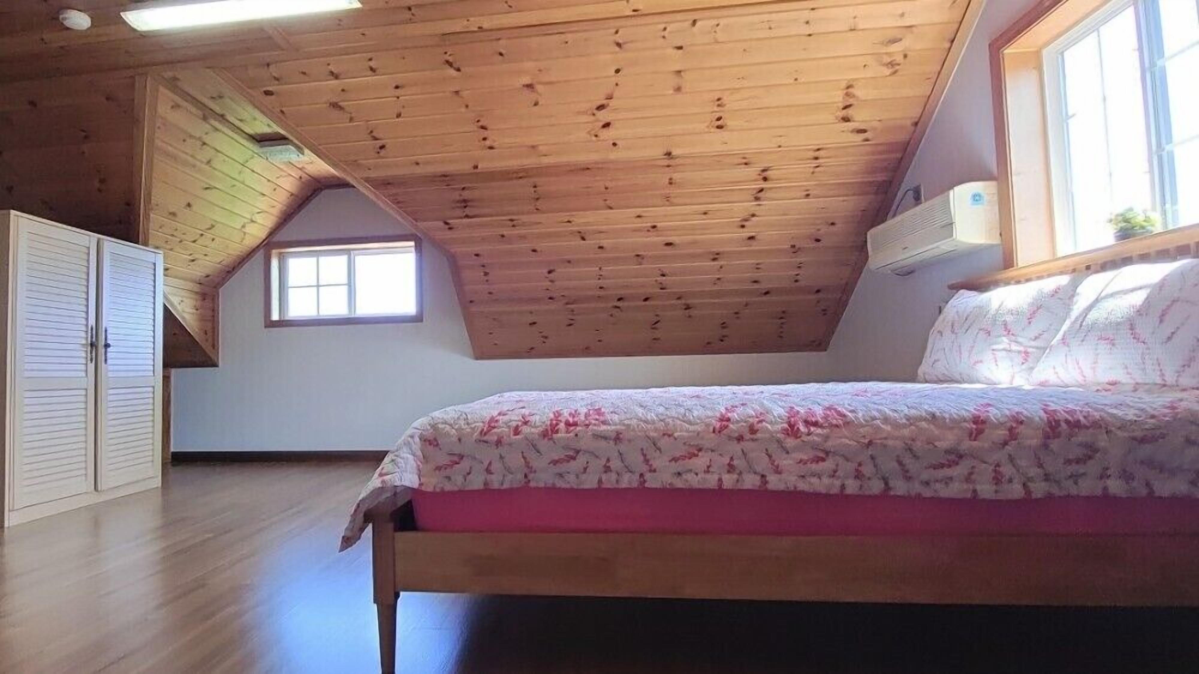 Une chambre avec un plafond en pente, des poutres en bois, un lit recouvert d’une couvre-lit à motifs floraux, une fenêtre et une armoire.