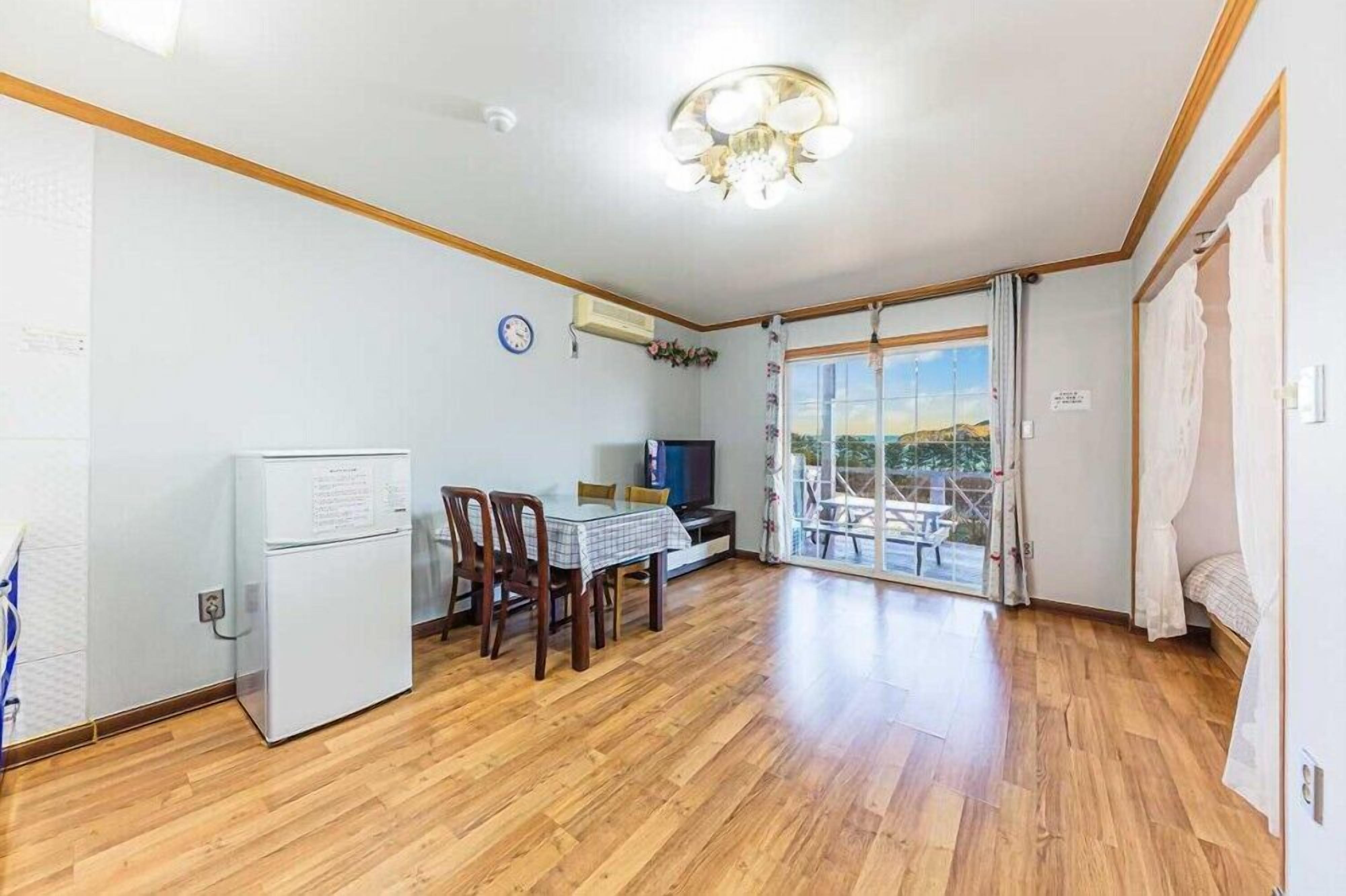 A spacious room with a wooden floor, a dining table with chairs, a refrigerator, a television, and a large window with a view of the outdoors.
