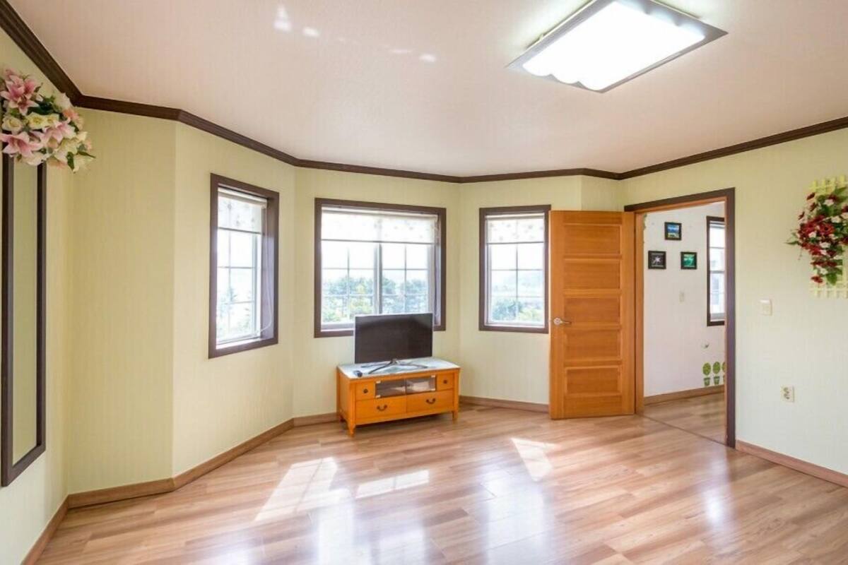 A spacious room with wooden flooring, a wooden cabinet, and a television. There are three windows providing natural light, and a door leading to another room.