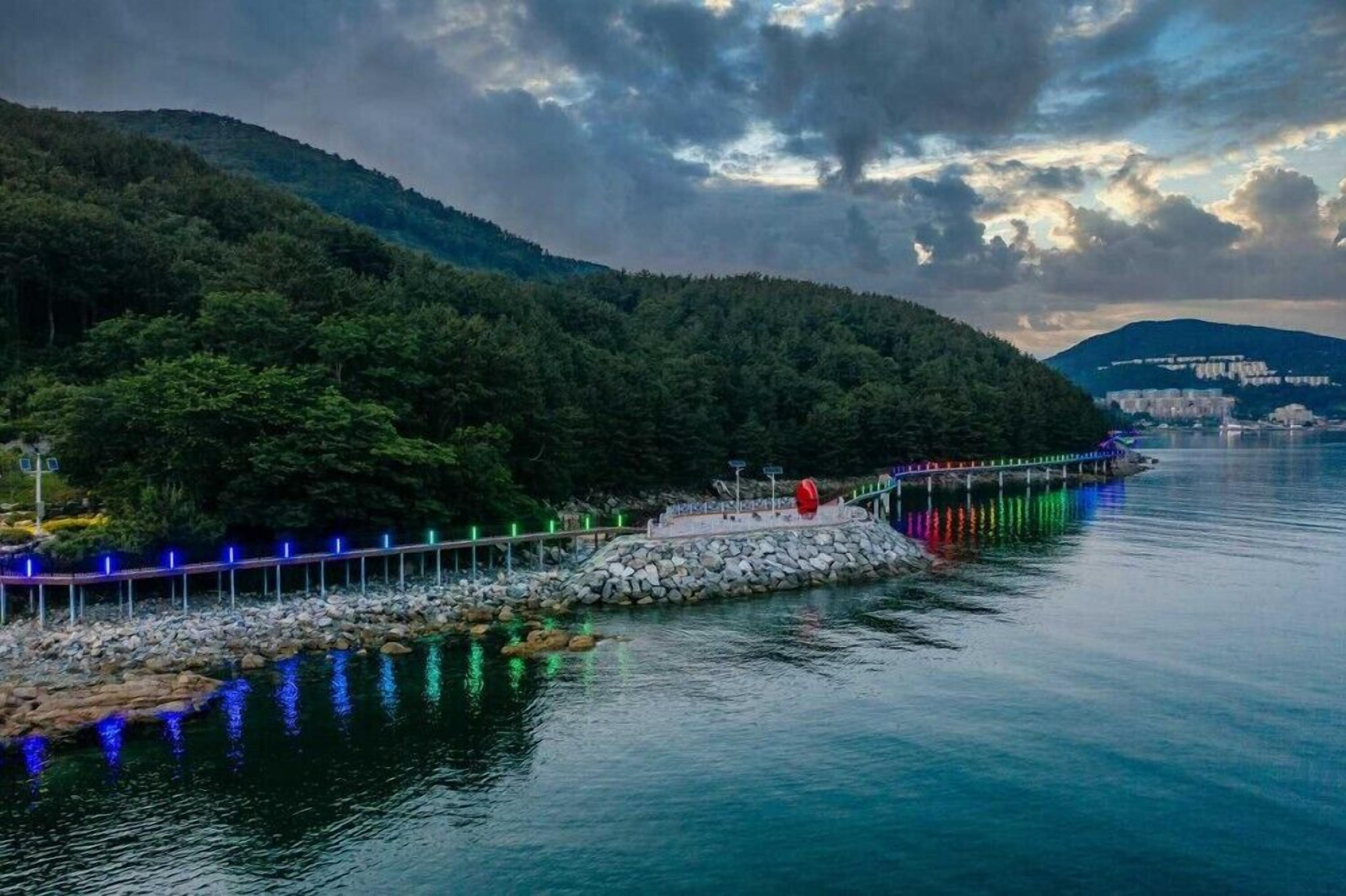 A waterfront promenade with a stone pier, lush greenery, and a cityscape in the background.