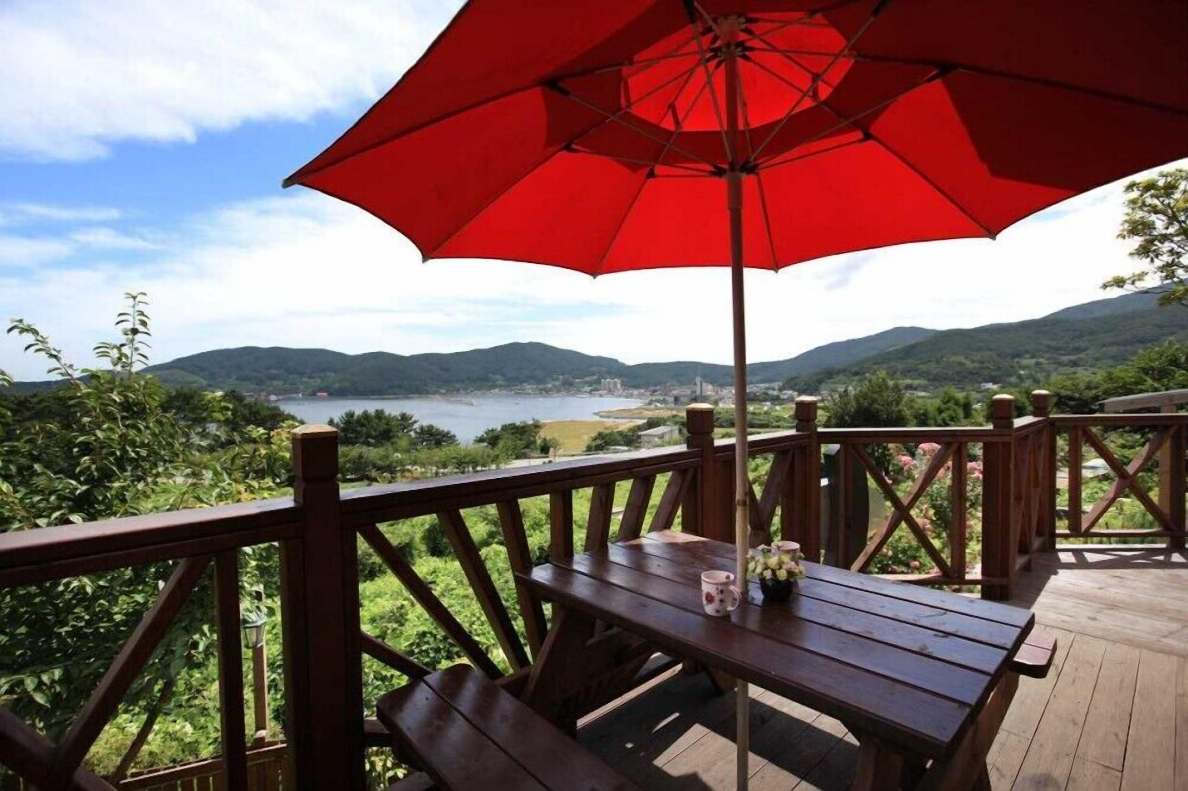 A wooden deck with a red umbrella, a table with a mug, and a scenic view of a lake and mountains.
