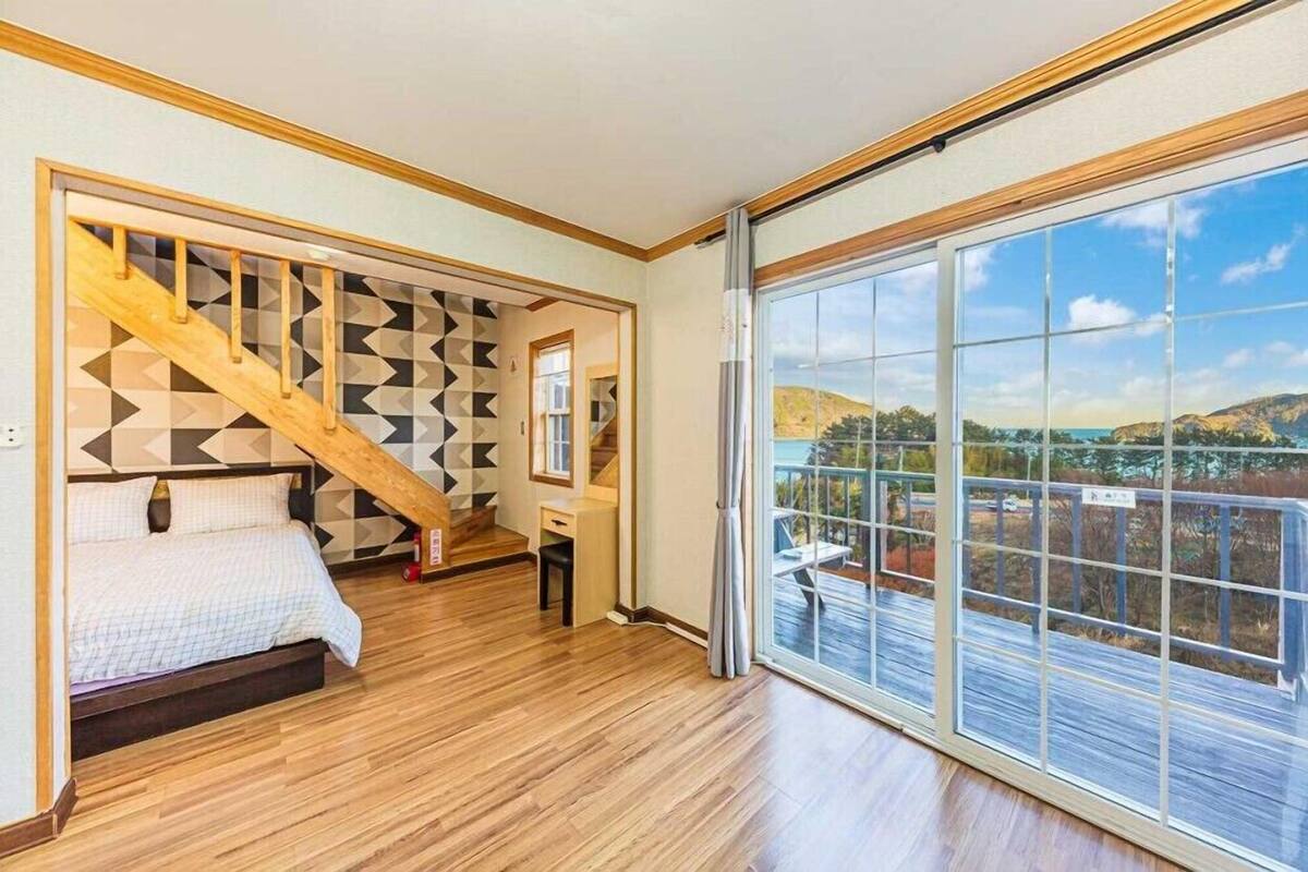 A bedroom with a wooden floor, a bed, a desk, and a large window with a view of hills.