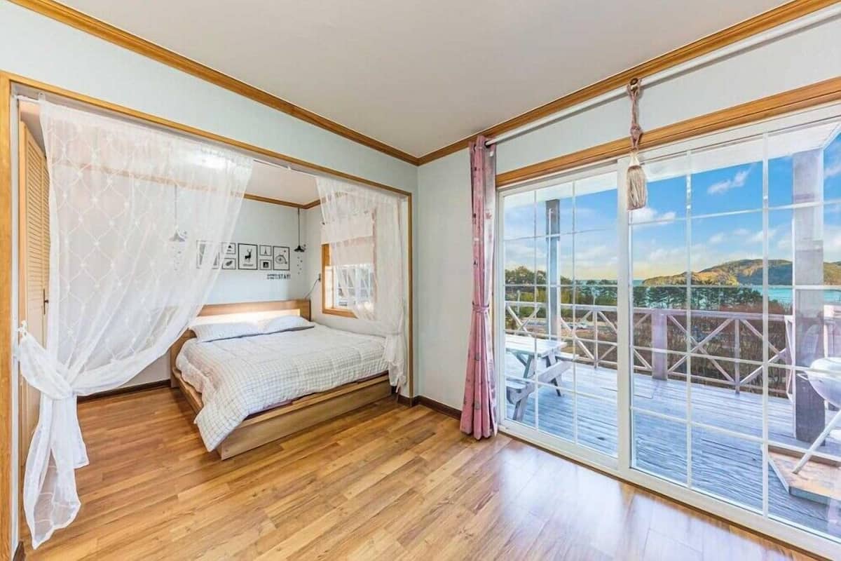 A bedroom with a bed, wooden flooring, a large window with a view of a deck and mountains, and a curtain.