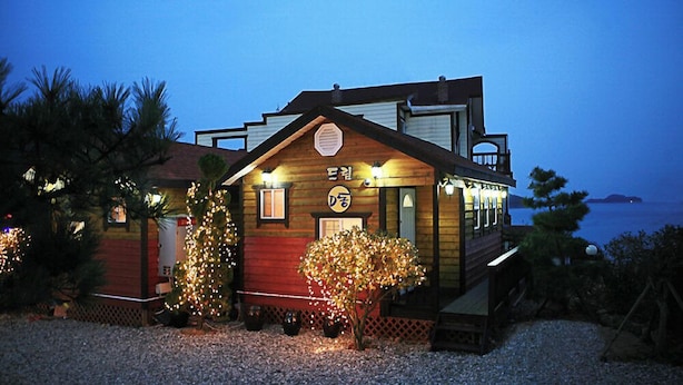 Una casa de madera con puerta roja y un balcón, rodeada de árboles y grava.
