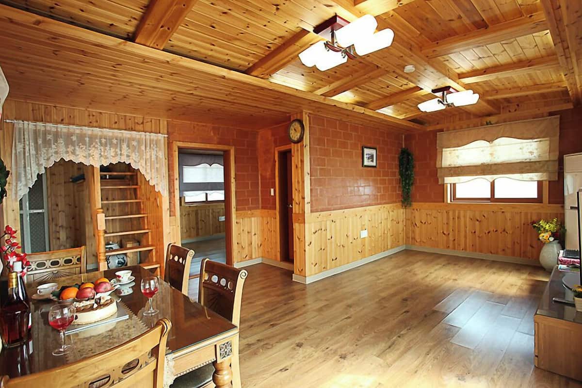 Interior de madera con una mesa de comedor preparada para dos personas, un reloj y una ventana con cortinas.