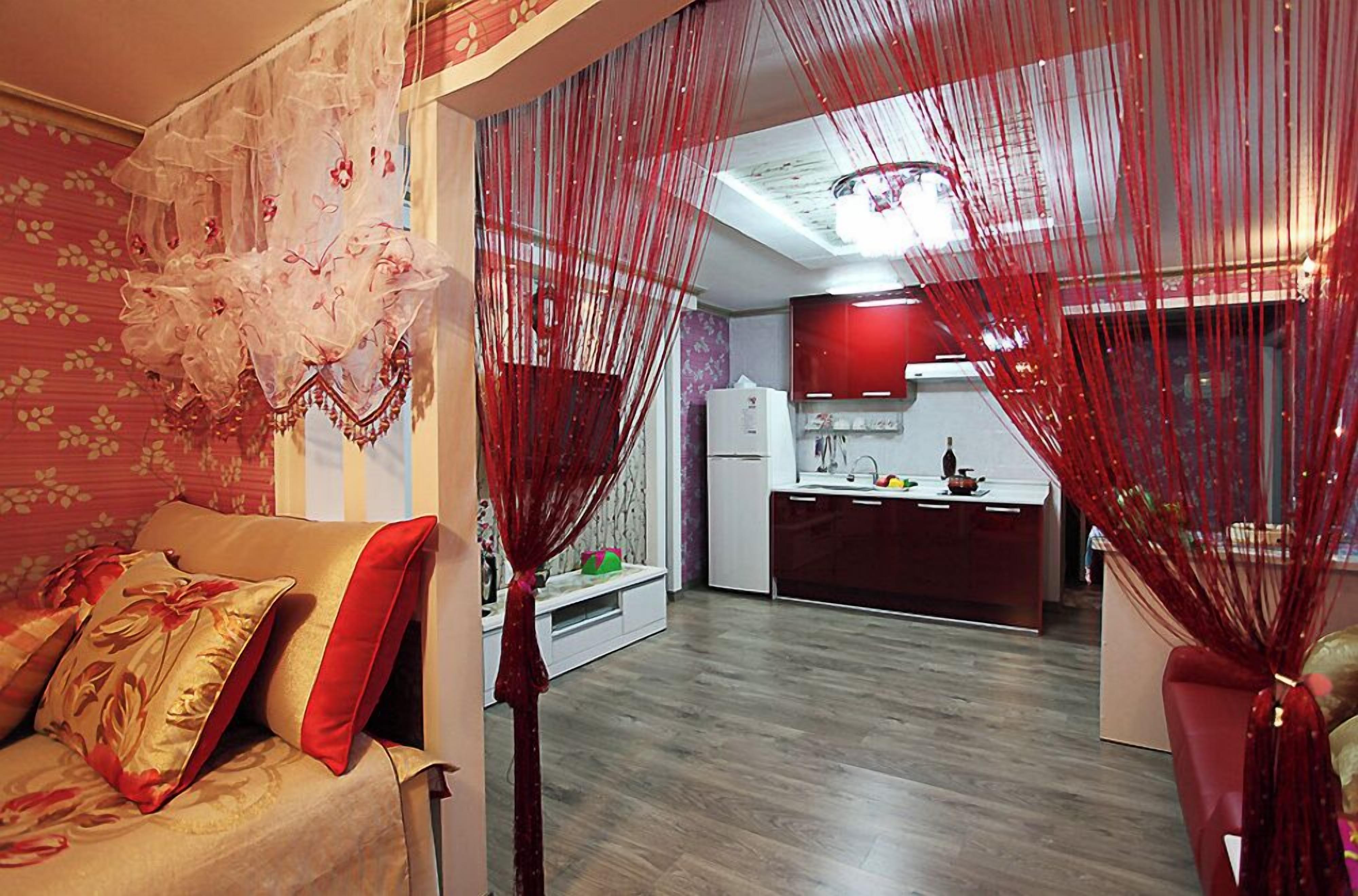 Una sala de estar con un sofá, una cocina al fondo y cortinas rojas.