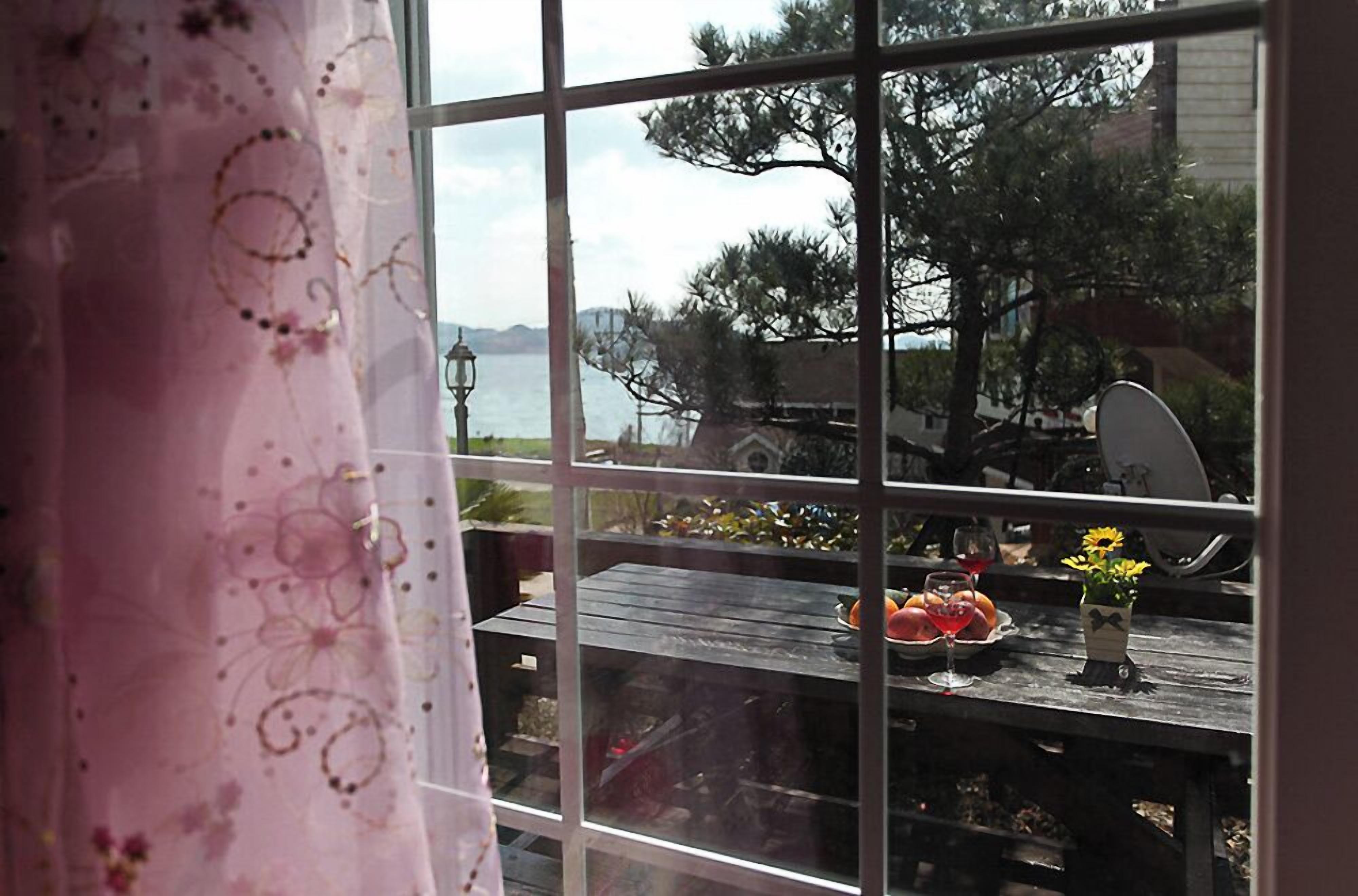 Une vue à travers une fenêtre ornée d’un rideau rose, une table en bois avec des fruits et un verre, et une vue sur un lac et des arbres.