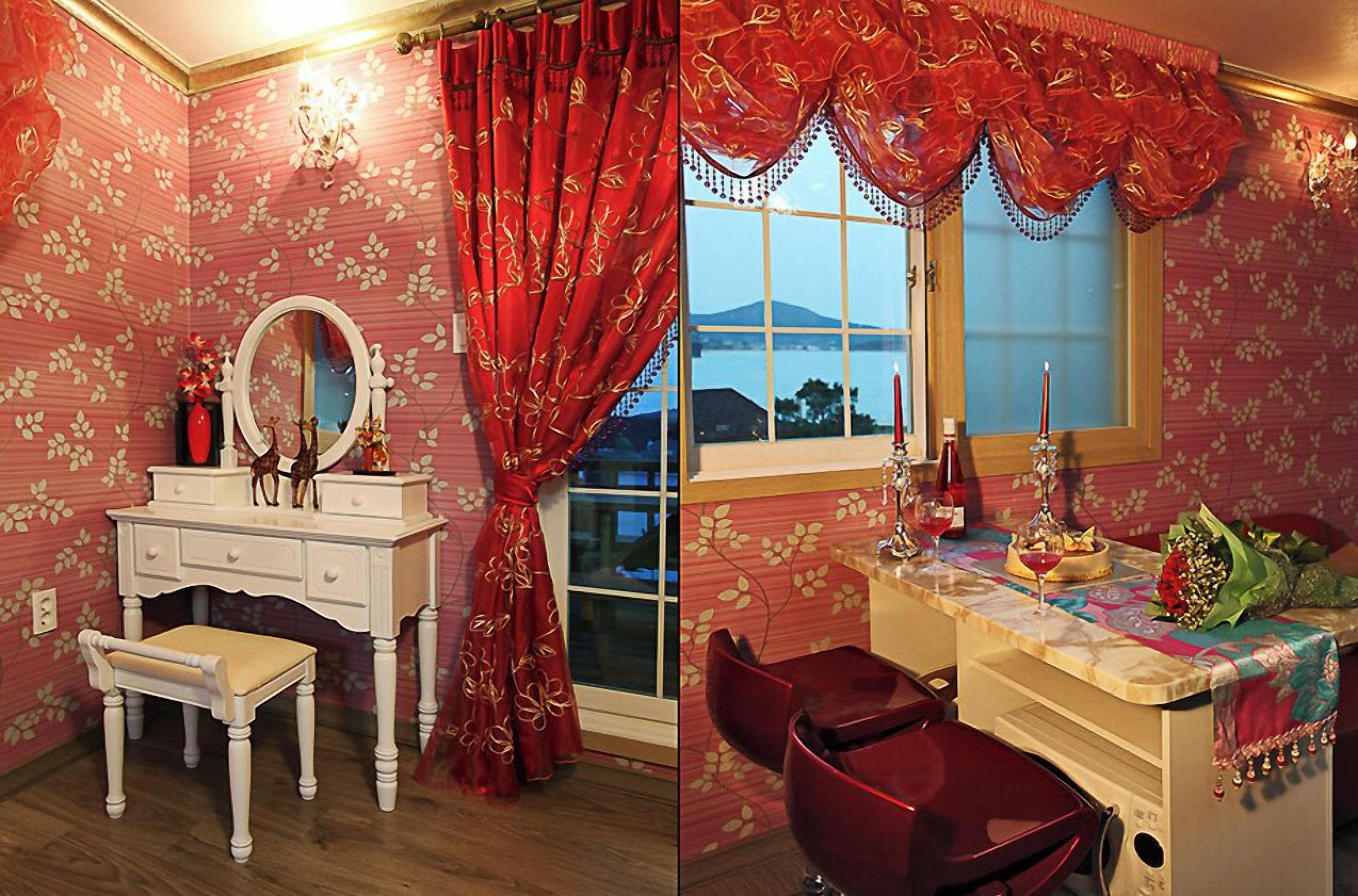 A room with floral wallpaper, a white vanity with a mirror, red curtains, and a view of a body of water through a window.