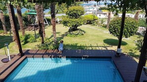 A swimming pool with a wooden deck, surrounded by greenery and a view of buildings in the background. - Haemaru pension (Geoje)