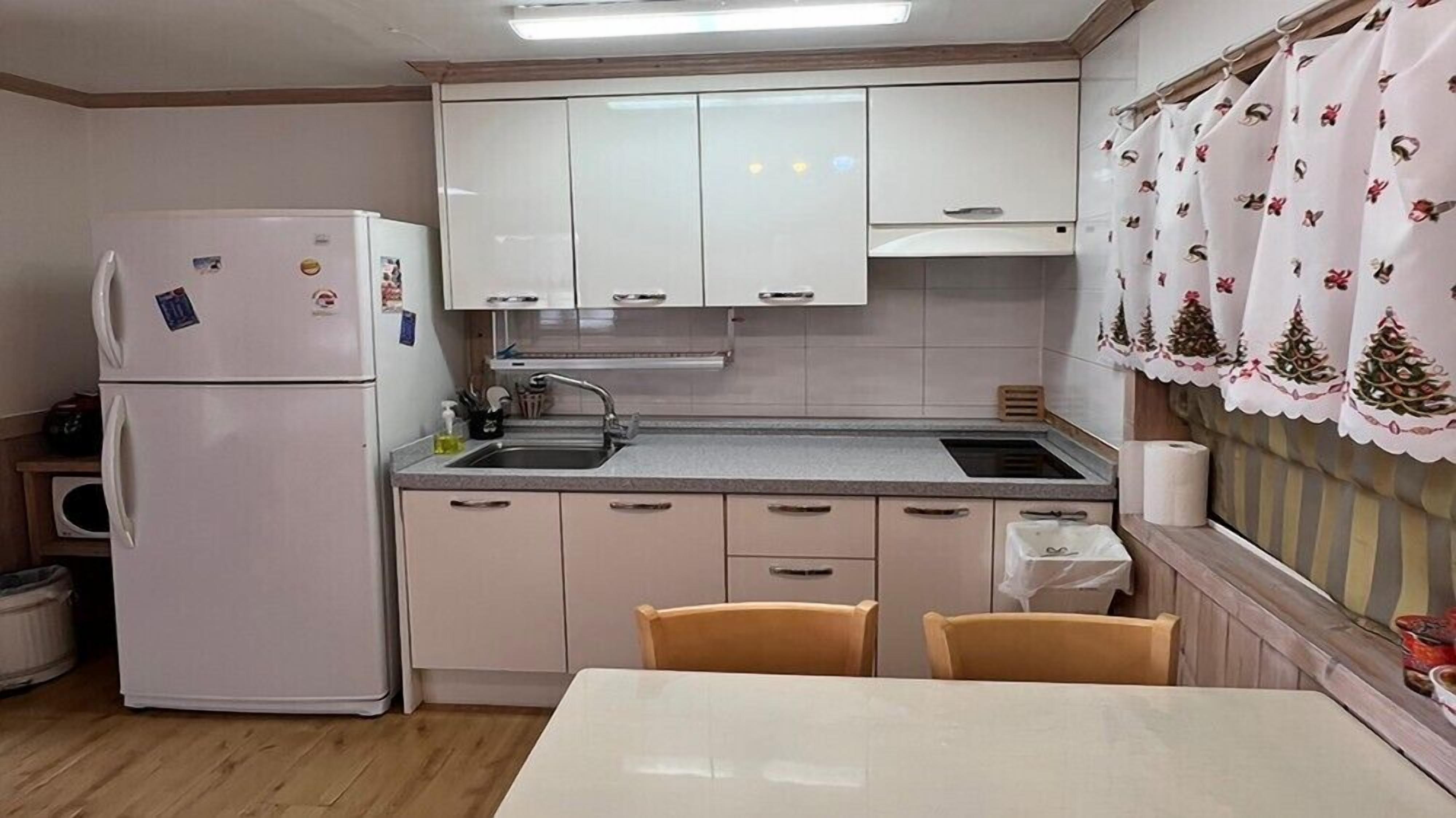 A compact kitchen with a refrigerator, sink, and wooden cabinets. There is a table with chairs and a window with curtains.