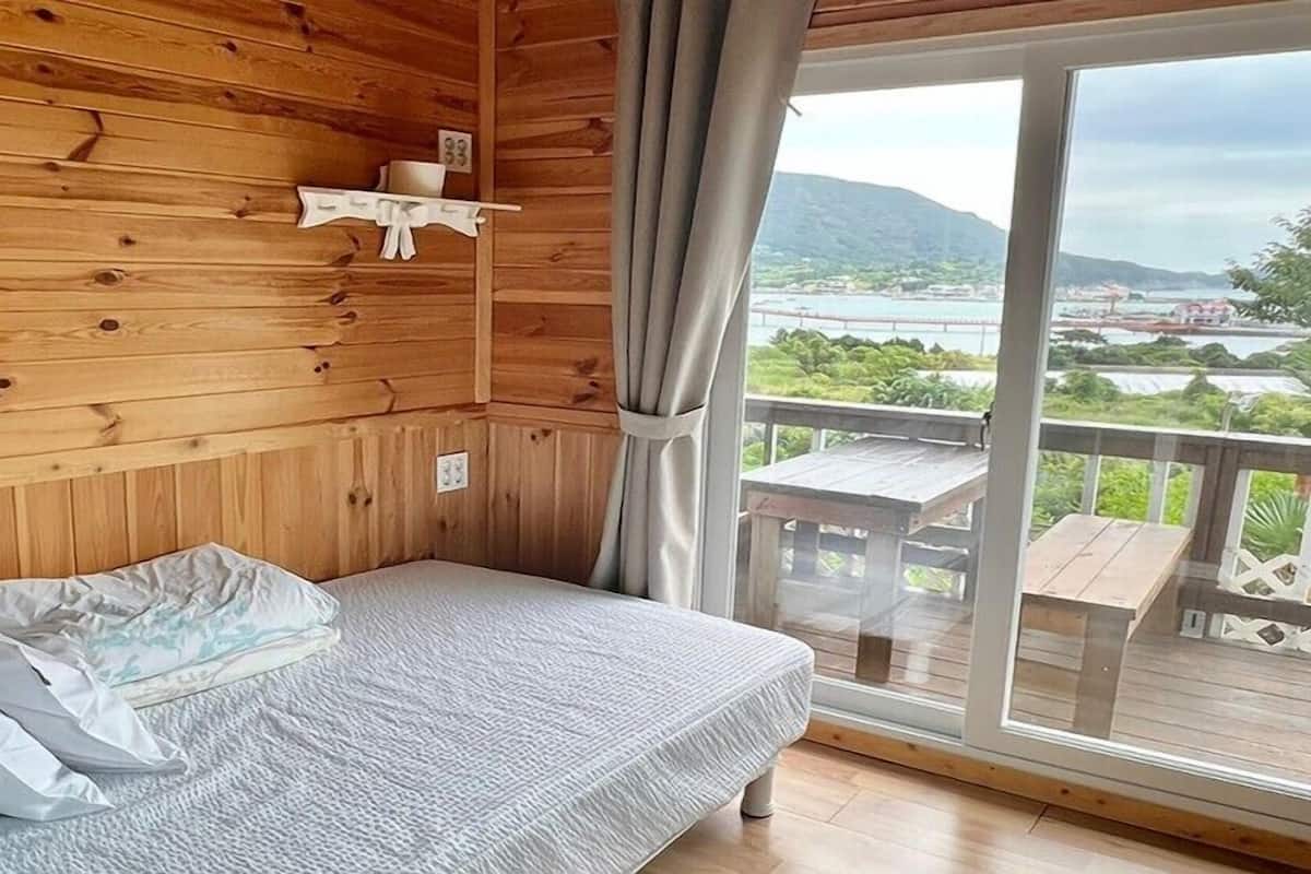 Una habitación tipo cabaña de madera con una cama, un estante pequeño y vista a un lago y montañas a través de un ventanal.