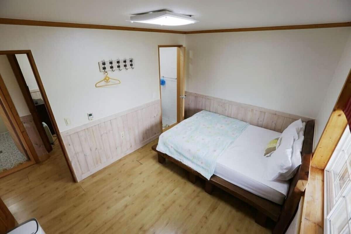 Un dormitorio con cama y suelo de madera, pared blanca con interruptor de luz colgante, y una puerta que da a otra habitación.