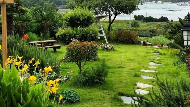 A garden with a stone pathway leading to a bench, surrounded by lush greenery and a view of the sea and mountains. - Haemaru pension (Geoje)