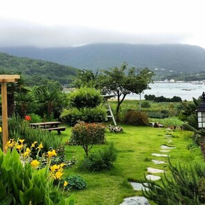 Un jardín con un sendero de piedra que lleva a un banco, rodeado de exuberante vegetación y vistas al mar y a las montañas.