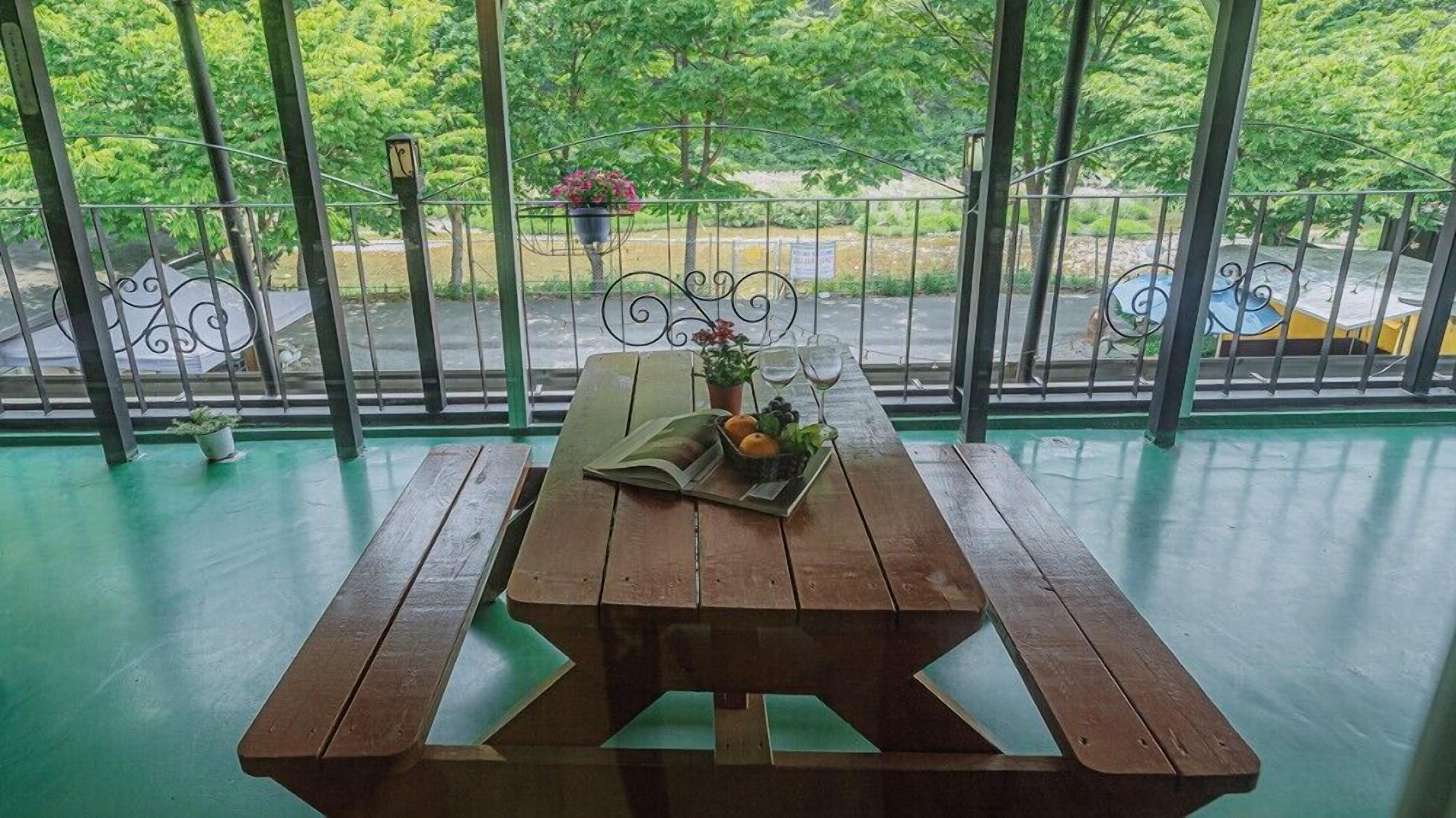 Une table de pique-nique en bois, sur laquelle sont posés un livre, des fruits et un verre de vin, est placée sur un sol vert, adossée à une rambarde métallique, avec vue sur des arbres et une route.