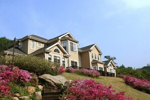 A large, multi-story house with a prominent front entrance, a driveway, and a landscaped garden with pink flowers.