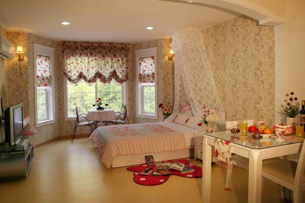 Un dormitorio con cama, una mesita de noche con lámpara, una silla, una pequeña mesa con lámpara y una ventana con cortinas florales.