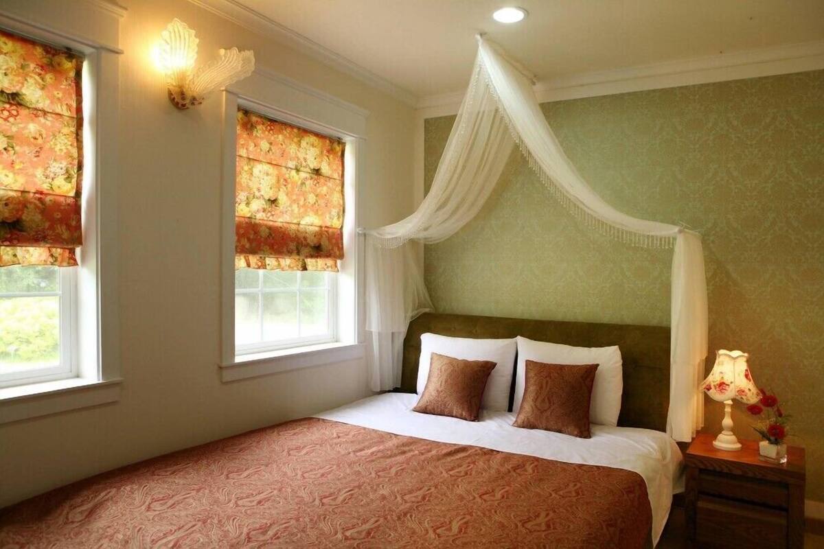 Una habitación con cama, mesita de noche, lámpara y ventana con cortinas.