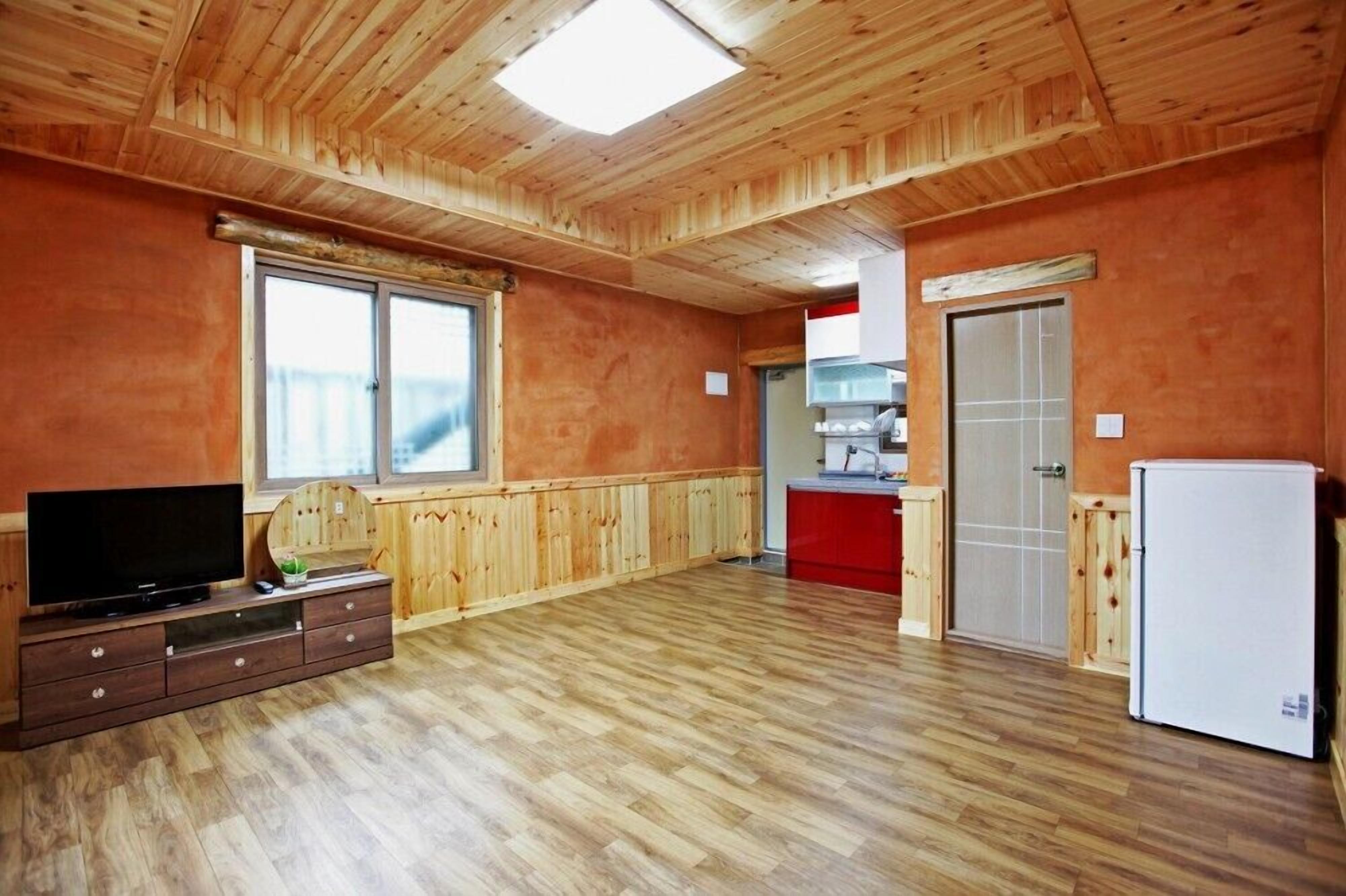 A spacious room with wooden flooring, a television on a wooden cabinet, a refrigerator, and a kitchen area with a red countertop.