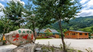 A rustic lodge with a large rock sign, wooden cabins, and a mountainous backdrop. - Utbeodeong Pension (Chuncheon)