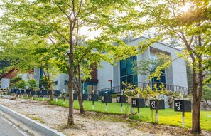 A modern building with a flat roof and large windows, surrounded by trees and a gravel path.