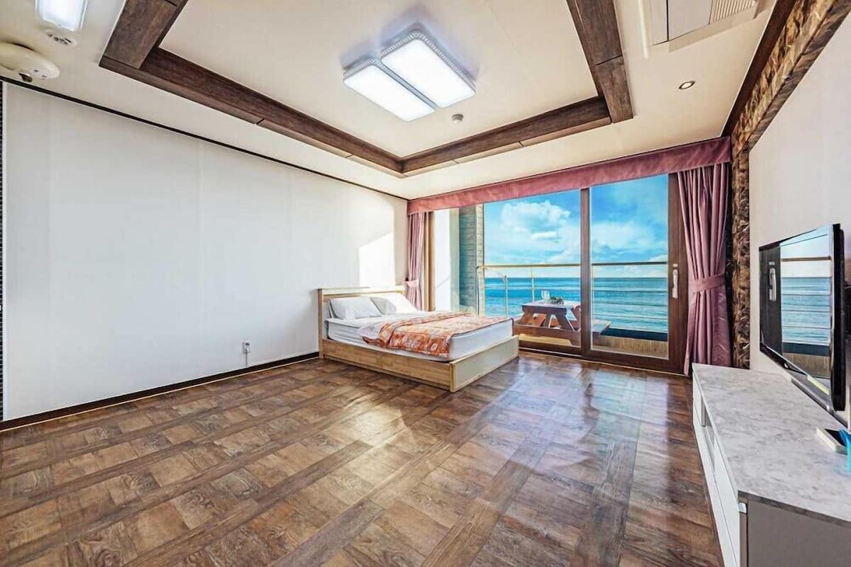 A bedroom with a wooden floor, a bed, a TV, and a large window with a view of the sea.
