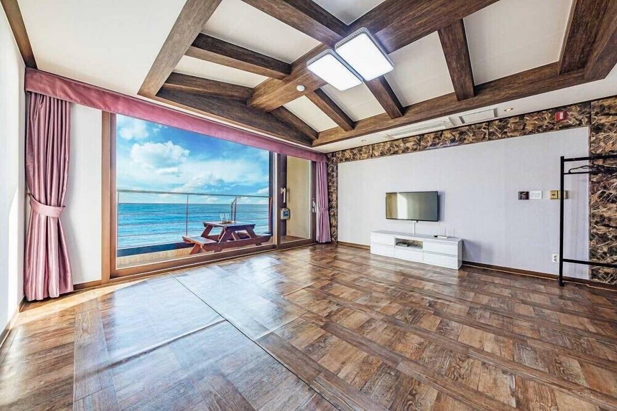 A spacious room with a wooden floor, a large window with a view of the sea, a flat-screen TV, and a white cabinet.