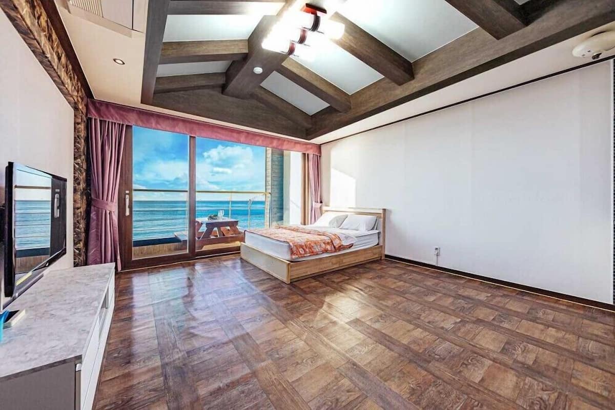 A bedroom with a wooden floor, a bed, a TV, and a view of the sea through large windows.