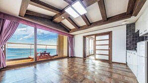 Une pièce spacieuse dotée d’un plancher en bois, de grandes portes-fenêtres coulissantes donnant sur un balcon avec vue sur la mer, et d’un coin cuisine avec des meubles blancs.