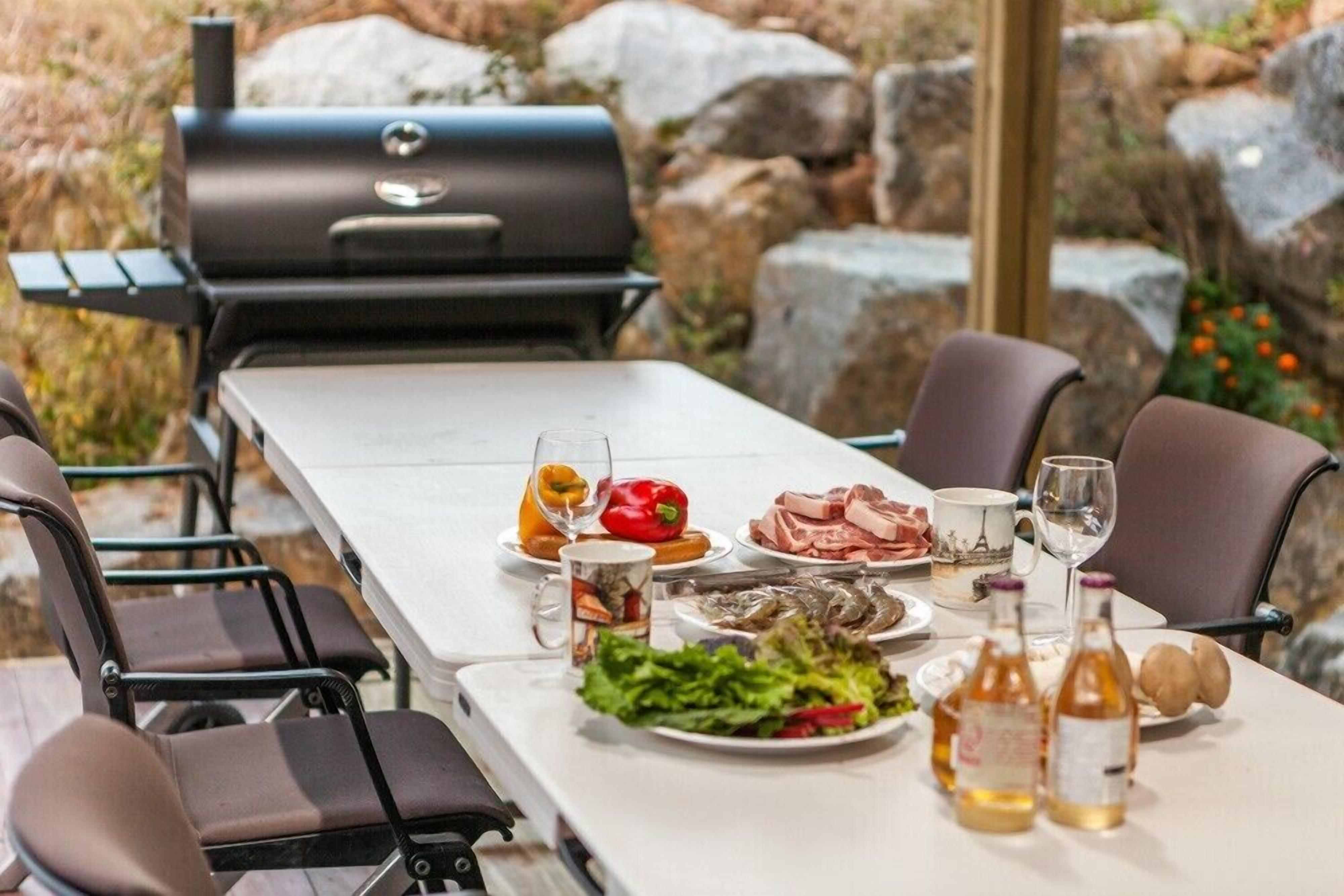 An outdoor dining area with a grill, chairs, and a table set with food and drinks.