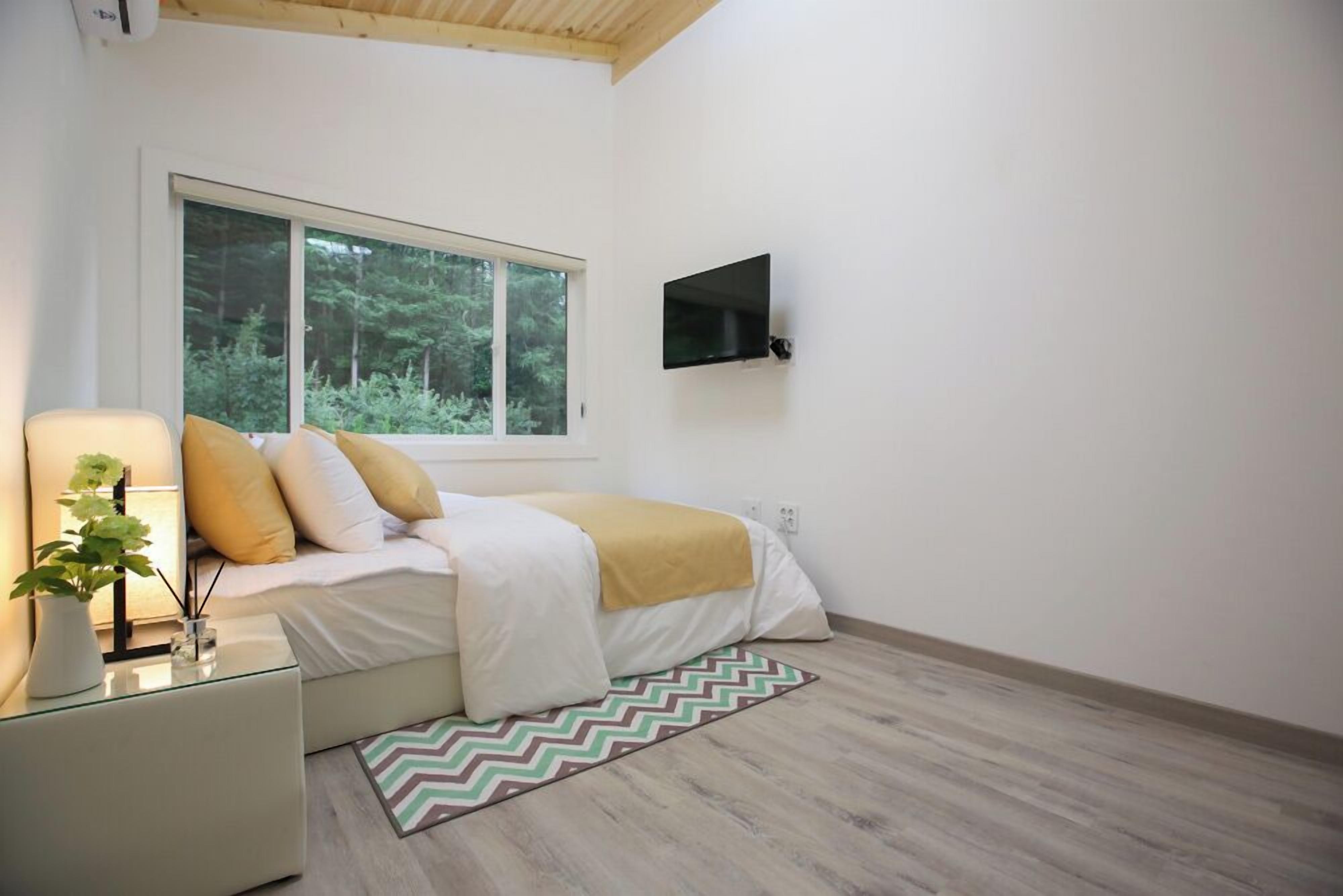 Un dormitorio moderno con cama, televisor montado en la pared, ventana con vista a árboles y mesita de noche con lámpara y planta.