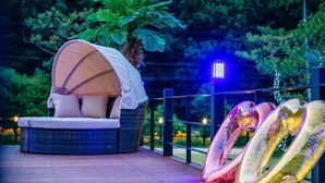 Un espacio circular para sentarse al aire libre con un banco acolchado y una marquesina, rodeado por una barandilla y un jardín con árboles e iluminación.