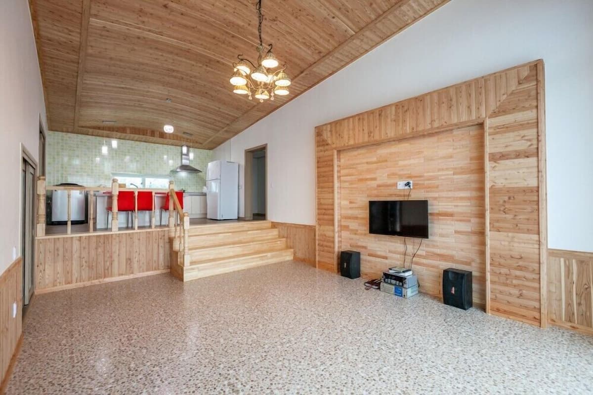 Un vaste espace de vie doté d’un plafond en bois, une cuisine à l’arrière-plan et un téléviseur fixé au mur.