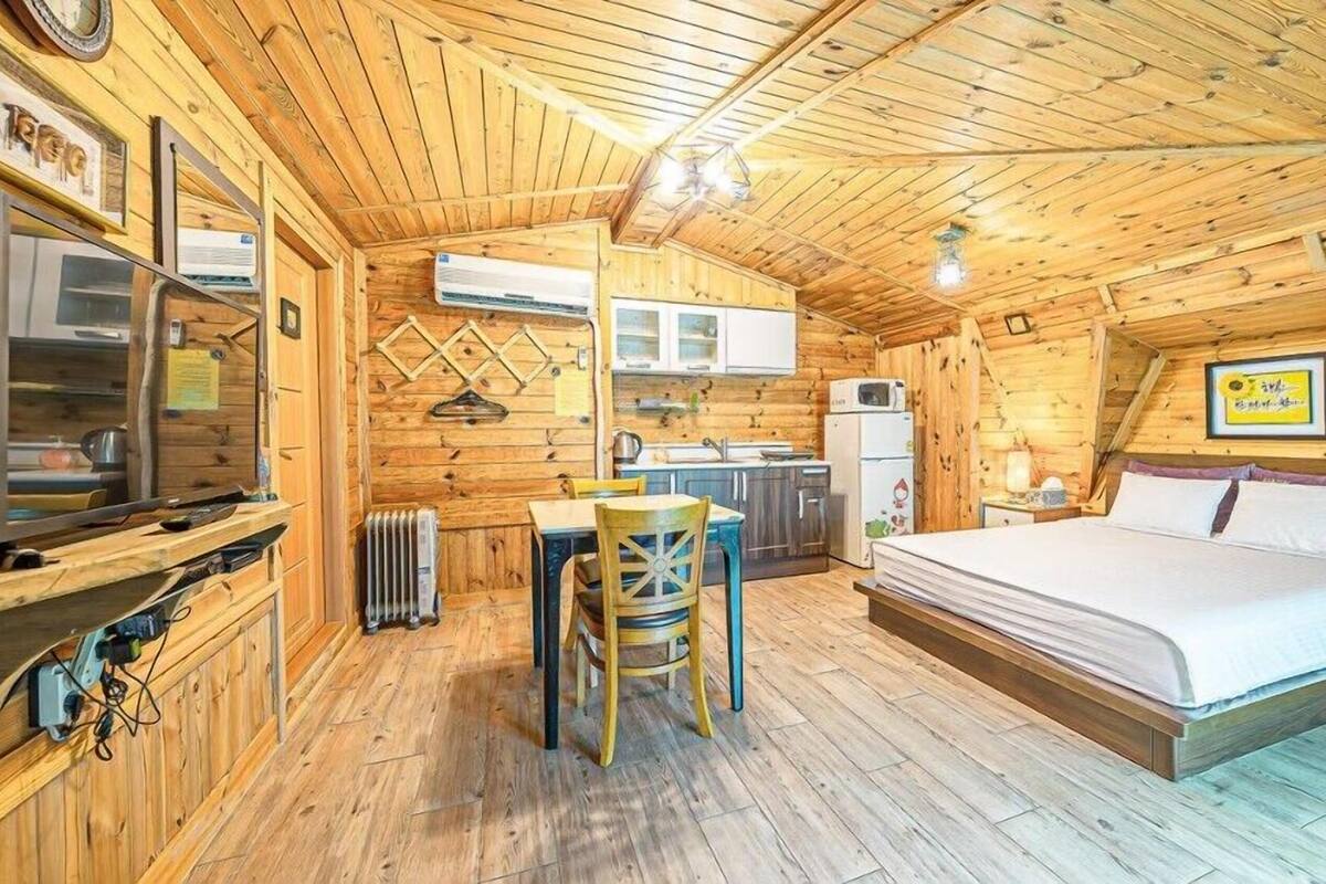 A cozy log cabin room with a bed, a dining table with chairs, a kitchen area with cabinets and a refrigerator, and a mounted TV.