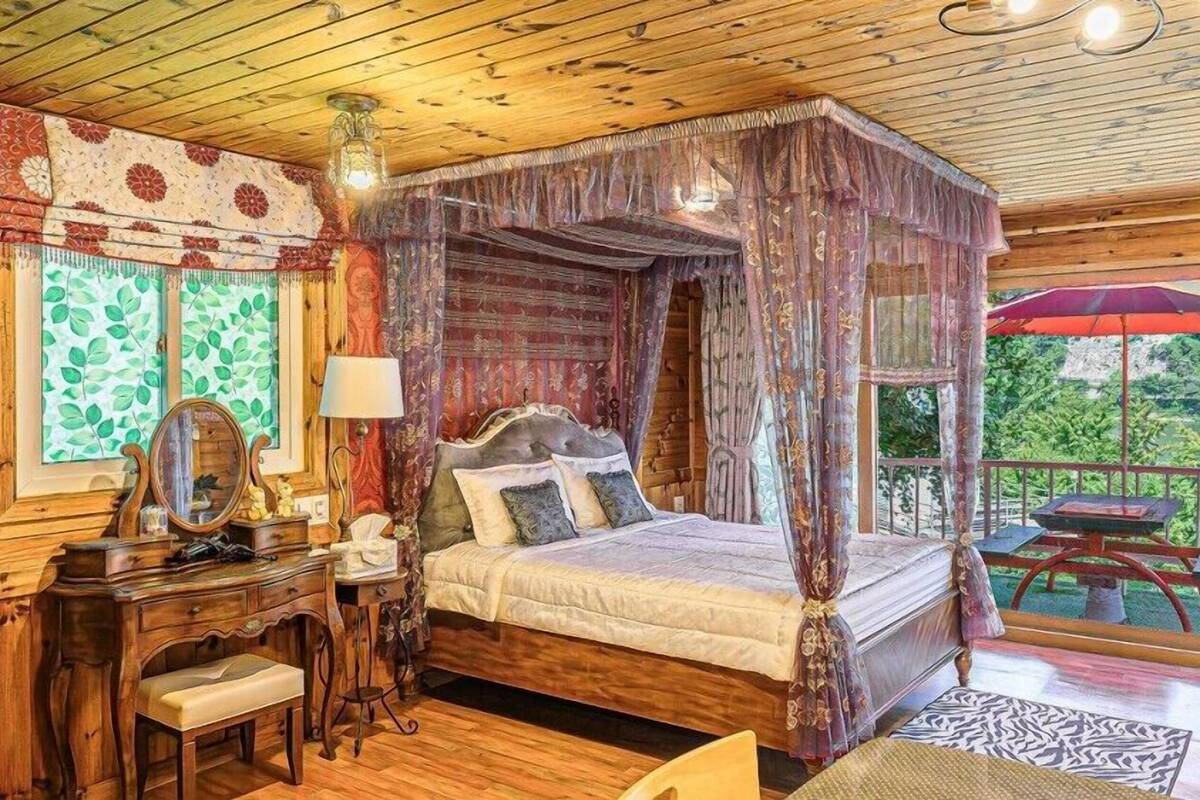 A four-poster bed with a canopy, a wooden desk with a mirror, and a view of the outdoors through large windows.