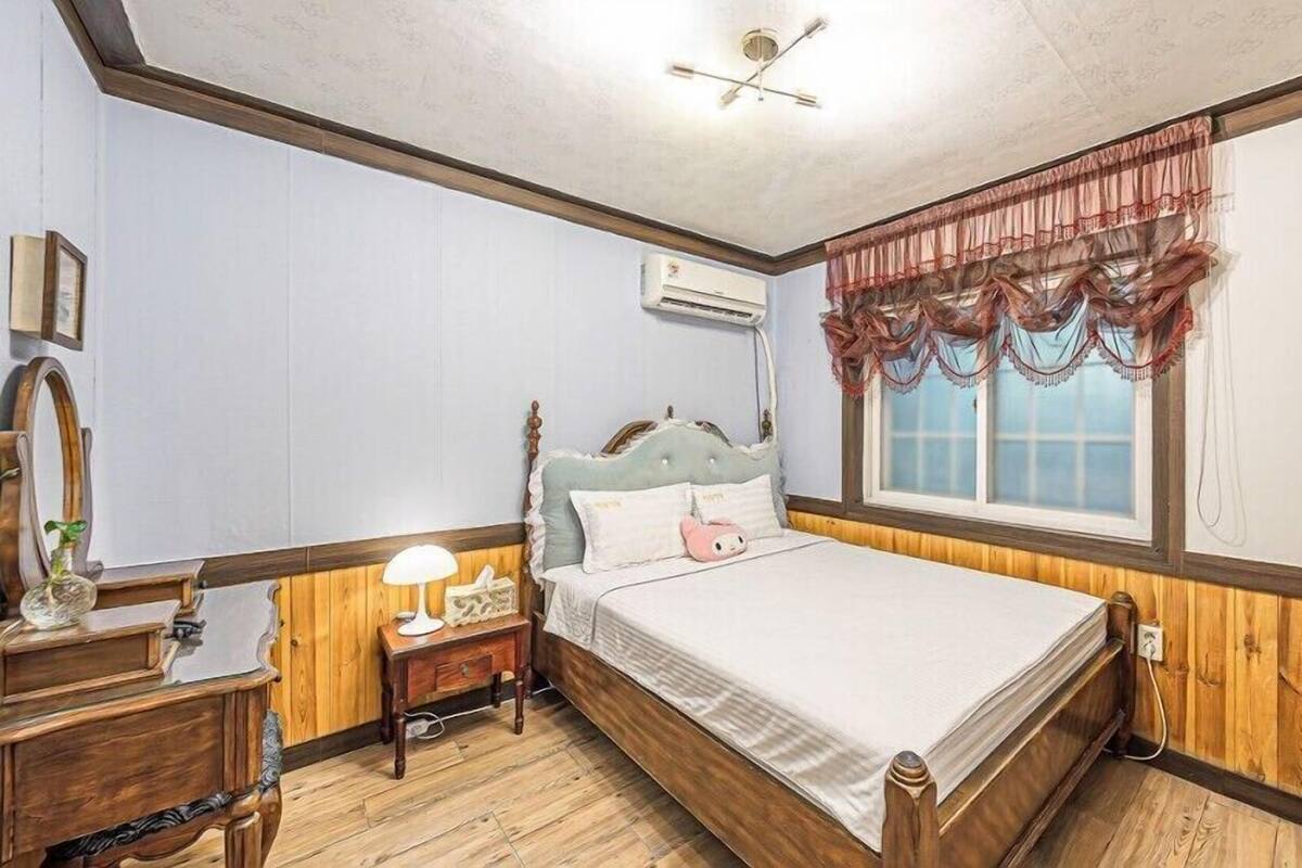 A bedroom with a wooden bed, a bedside table with a lamp, a mirror, and an air conditioning unit on the wall.