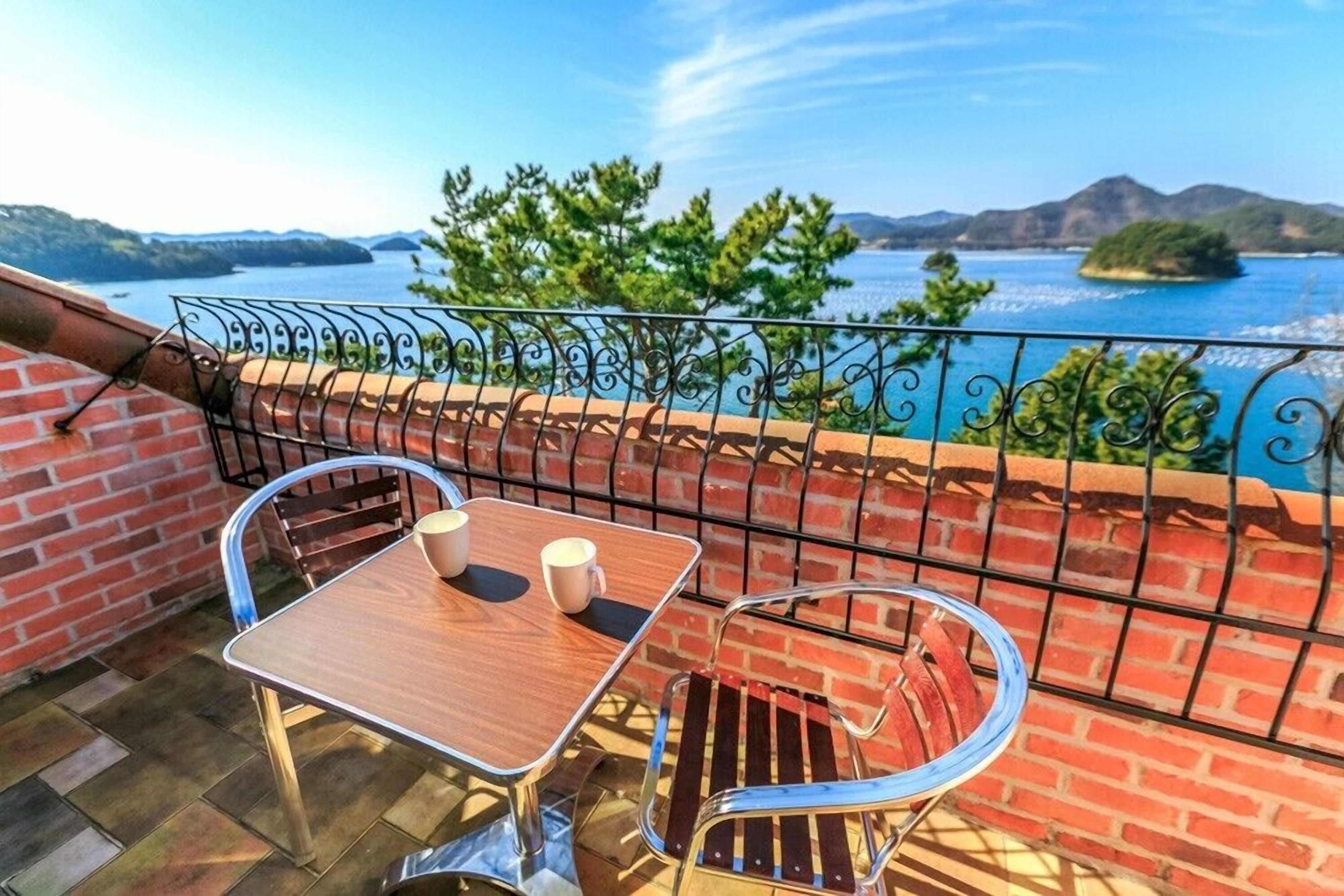 A balcony with a table and chairs, a brick wall, and a view of the sea and hills.