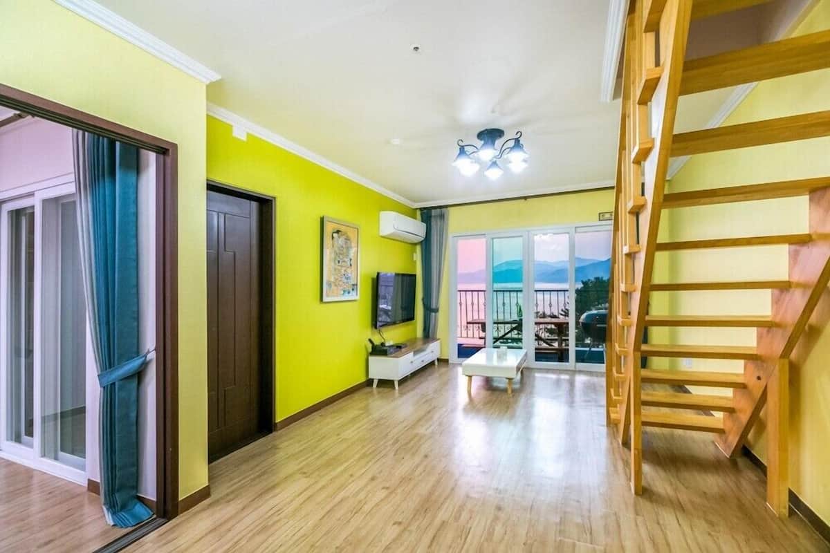 Un salón luminoso con una escalera de madera, un televisor y vistas a través de un ventanal.