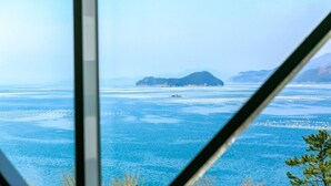 A view through a window of a coastal area with calm waters, a small island, and distant mountains. - Beach castle hotel & resort (Tongyeong)