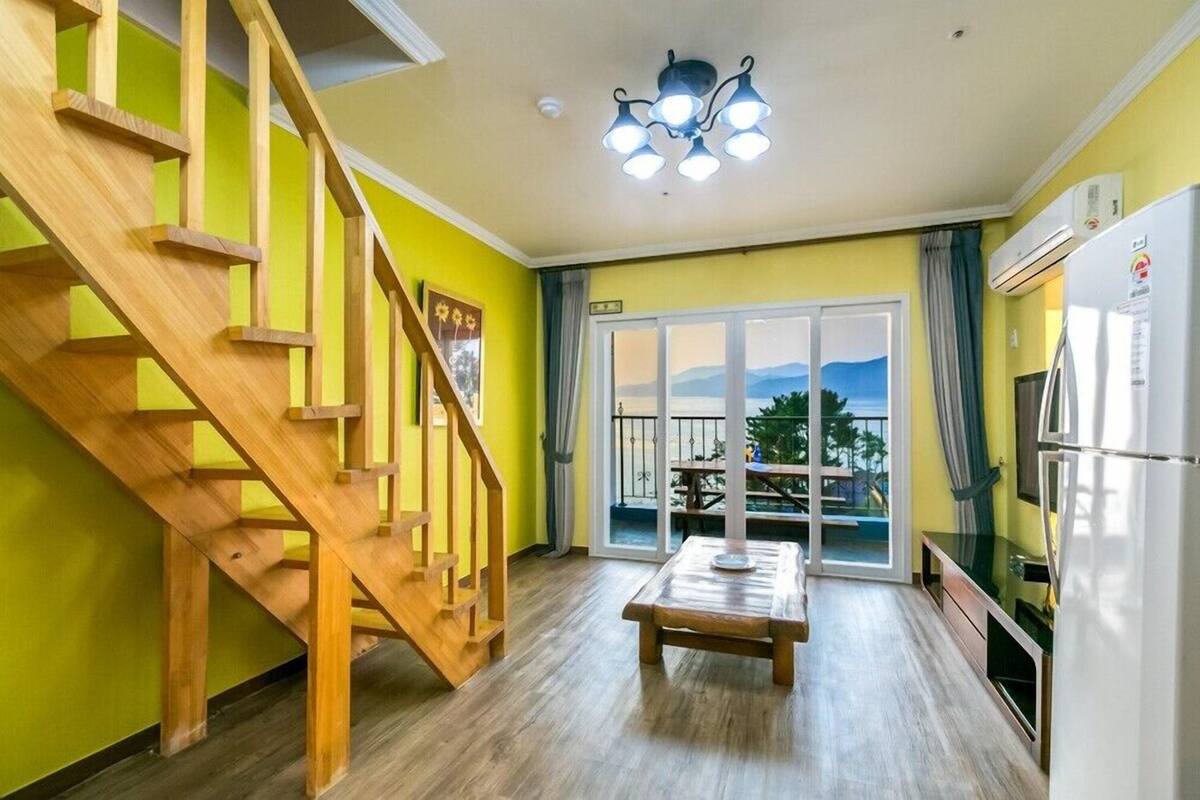 Una habitación con escalera de madera, una mesa de comedor, un refrigerador y vista a través de un ventanal.