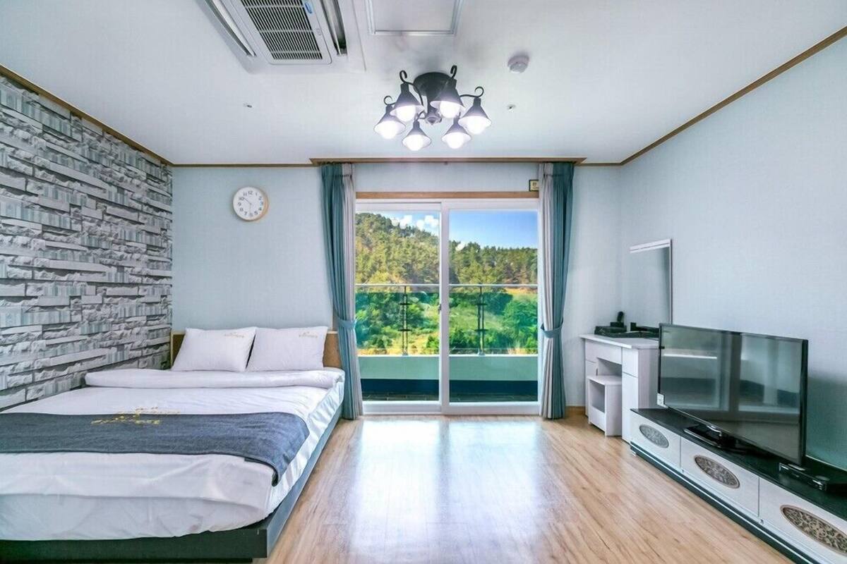 Un dormitorio con cama, televisor, ventana con vista a la vegetación y pared de piedra.