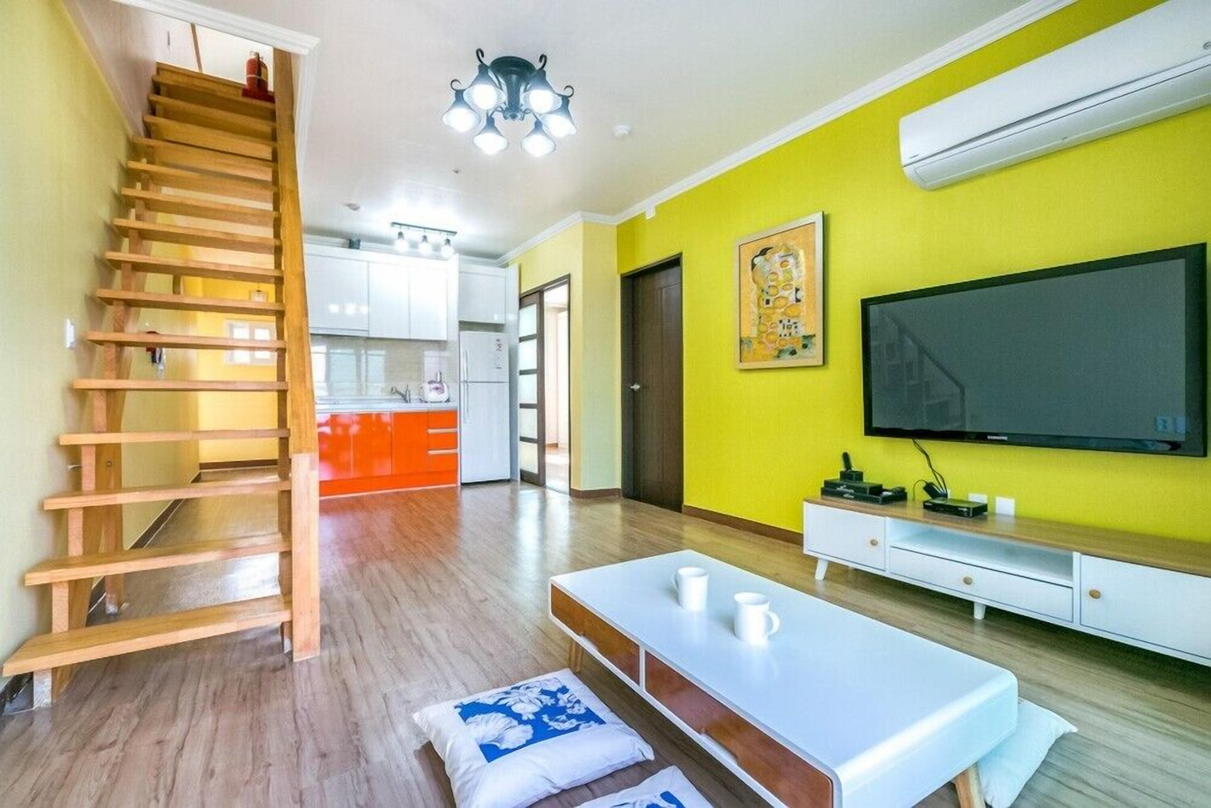 A modern living room with a wooden staircase, a flat-screen TV, a white coffee table, and a kitchen in the background.