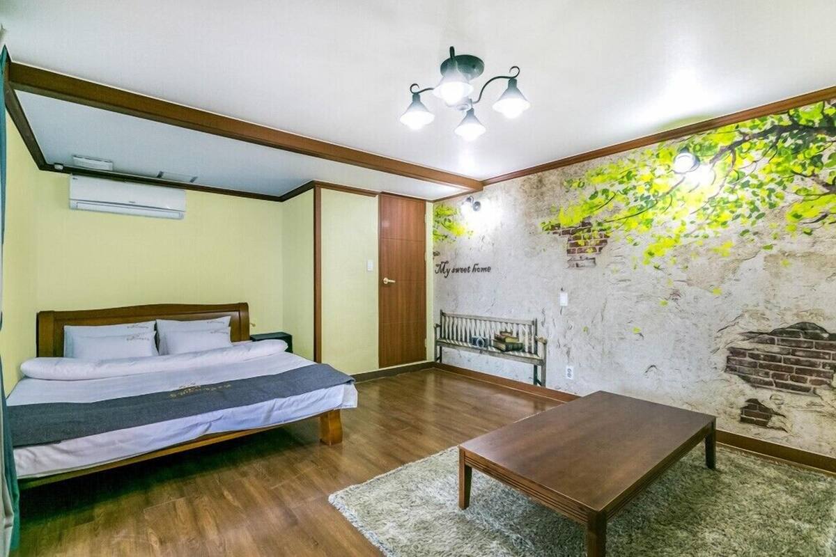Un dormitorio con cama, una mesa de madera, un banco, un mural en la pared y una lámpara en el techo.